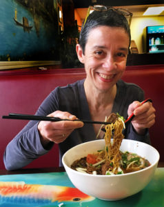smiling cropped hair woman eating pho noodles with chopsticks